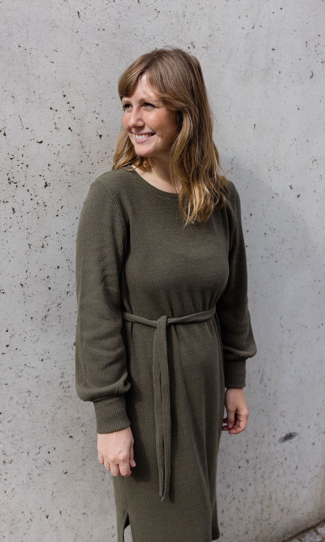 Strickkleid Jules in grün olive mit Stiefeln an heller Wand angelehnt Blick seitlich
