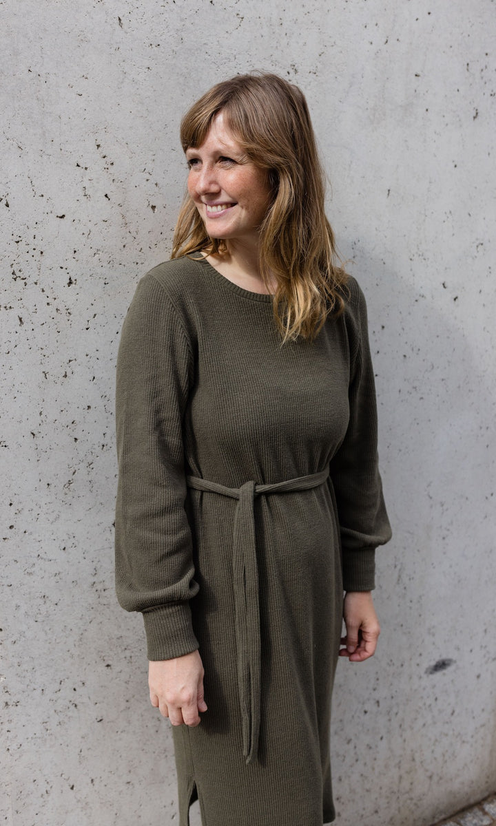 Strickkleid Jules in grün olive mit Stiefeln an heller Wand angelehnt Blick seitlich