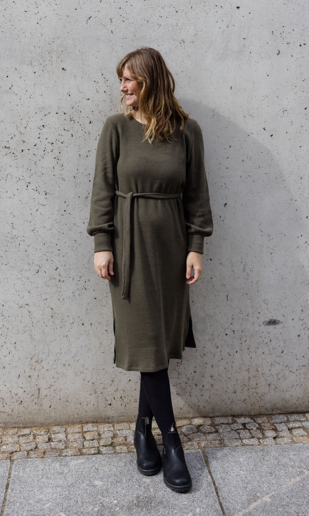 Strickkleid Jules in grün olive mit Stiefeln frontal vor Wand Blick seitlich