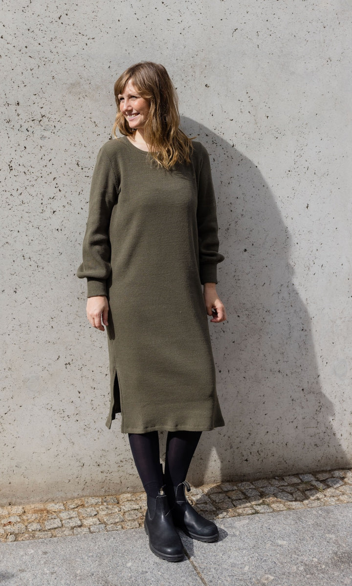 Strickkleid Jules in grün olive mit Stiefeln frontal vor Wand Blick seitlich in Sonne