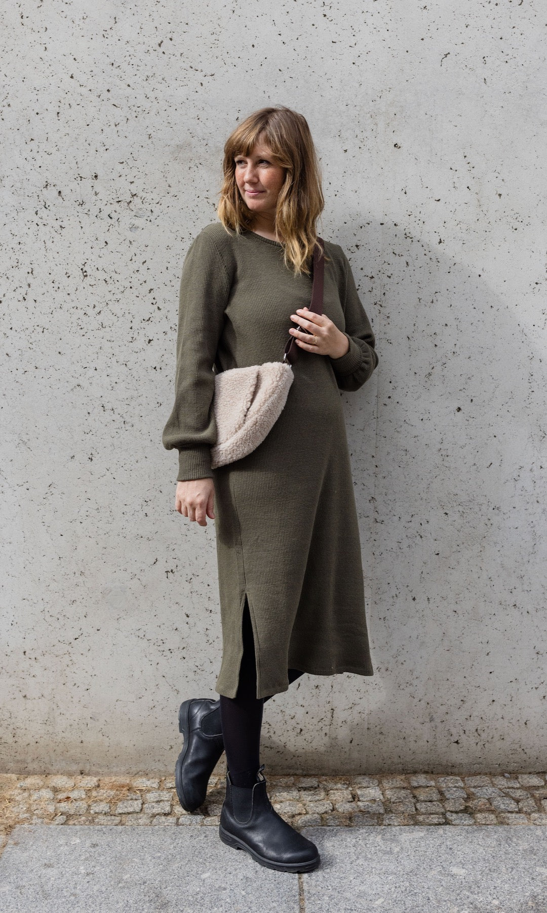 Strickkleid Jules in grün olive mit Stiefeln und Tasche seitlich vor heller Wand