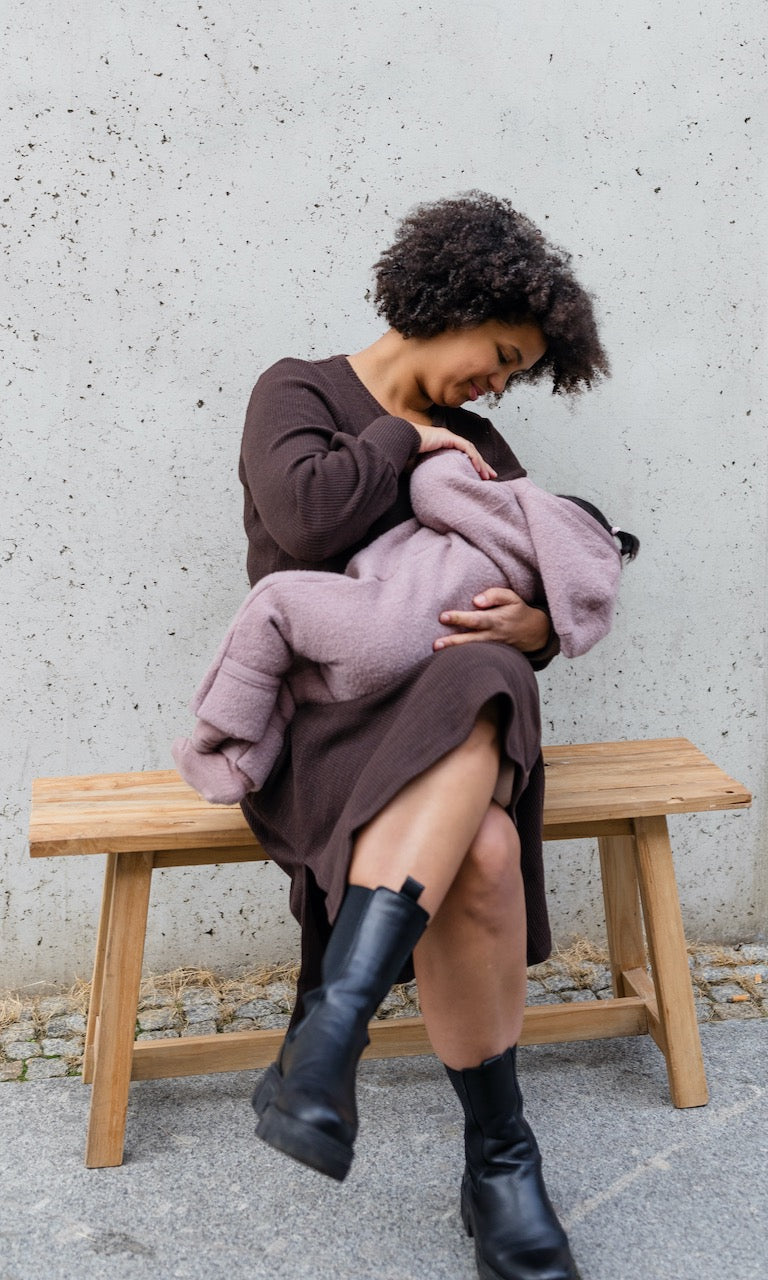Strickkleid Jules mit seitlicher Stillfunktion in braunem Strick mit schwarzen Stiefeln sitzend auf Bank beim Stillen mit Baby 