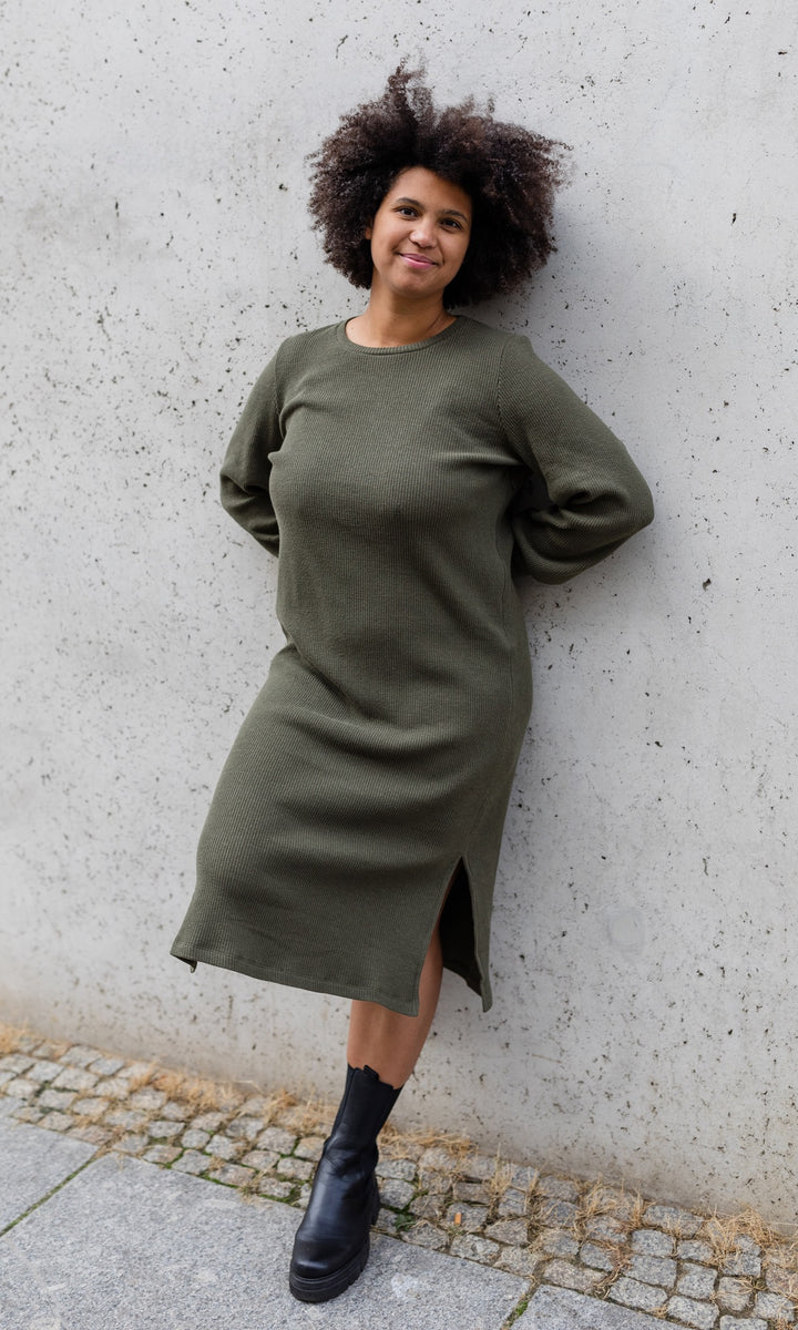 Strickkleid mit seitlicher Stillfunktion Jules in grün olive mit Stiefeln an heller Wand angelehnt