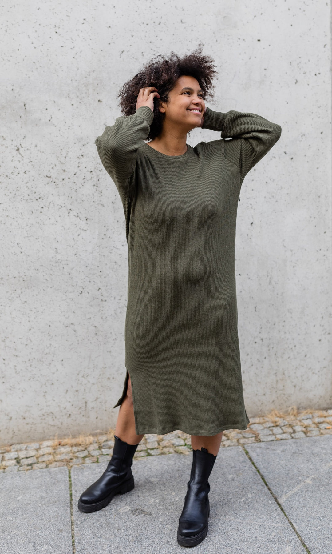 Strickkleid Jules in grün olive mit seitlicher Stillfunktion mit Stiefeln frontal vor heller Wand Blick seitlich
