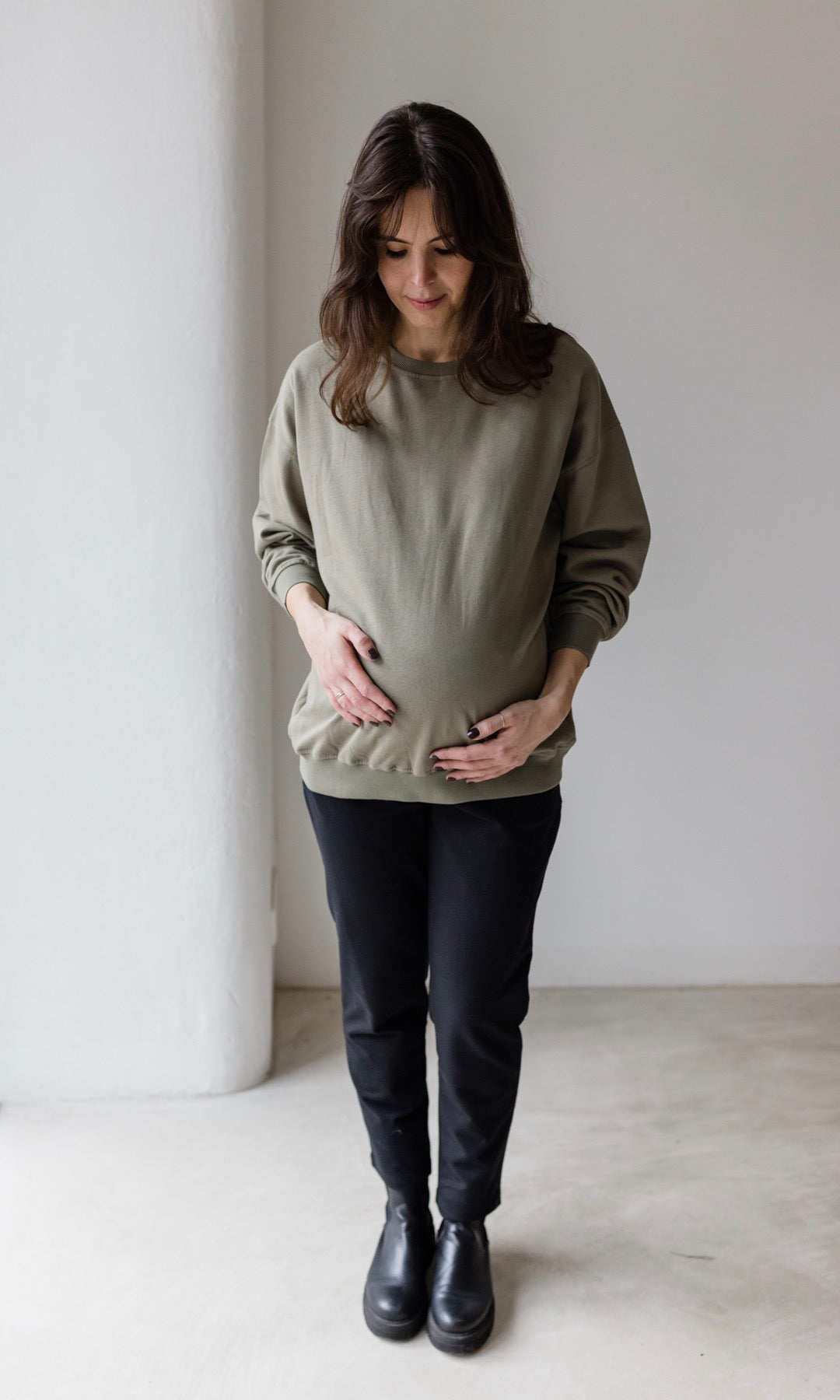 Vorderansicht Umstandshose Mom in schwarz kombiniert mit oliv grünem oversized Pullover und schwarzen Schuhen.