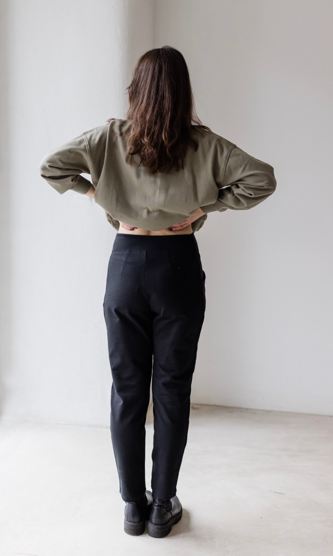Rückansicht Umstandshose Mom in schwarz kombiniert mit oliv grünem oversized Pullover und schwarzen Schuhen.