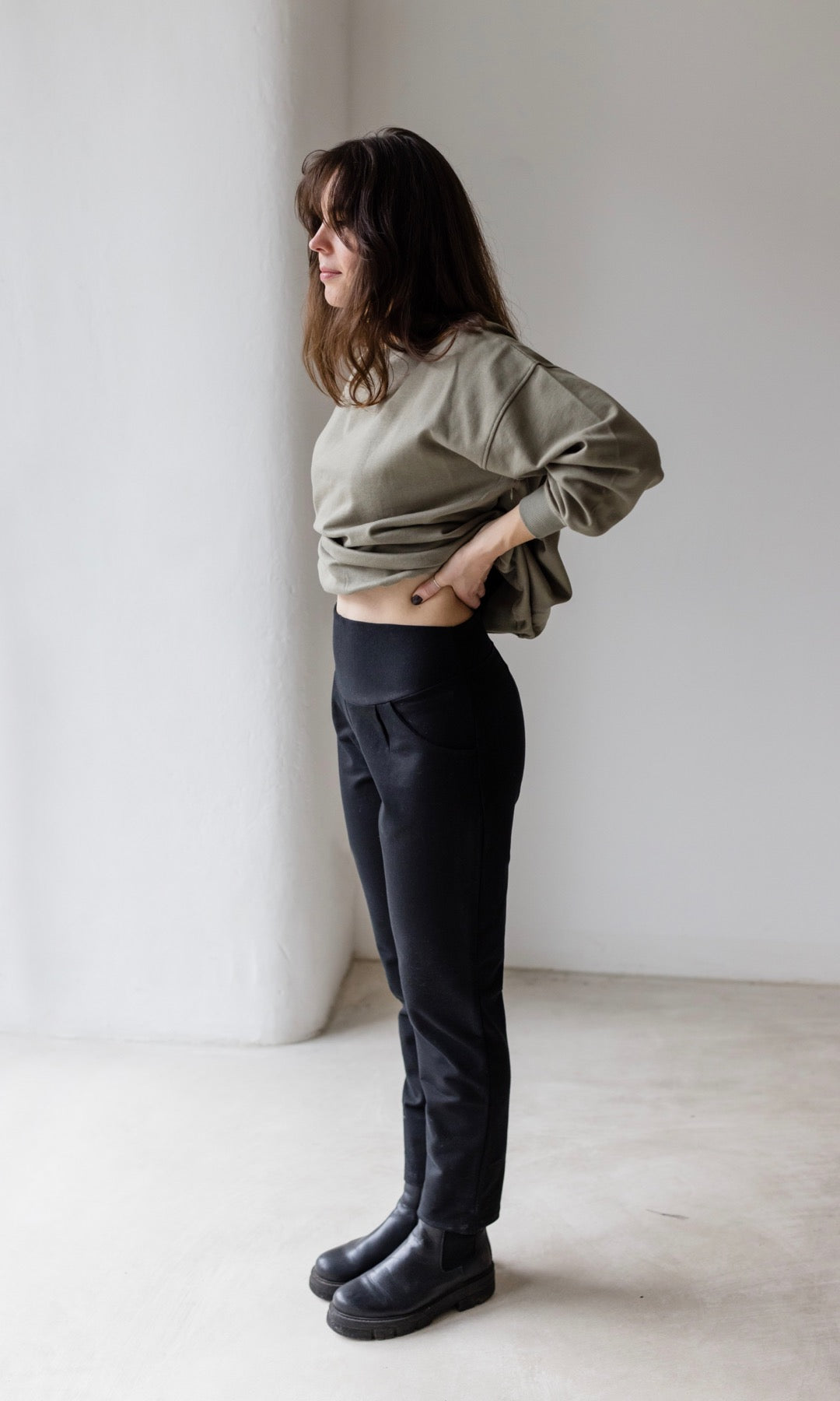 Seitliche Ansicht der Mom Umstandshose in schwarz mit Detailaufnahme Bauchbund aus Baumwolle. Kombiniert mit oliv grünem Pullover und schwarzen Schuhen.