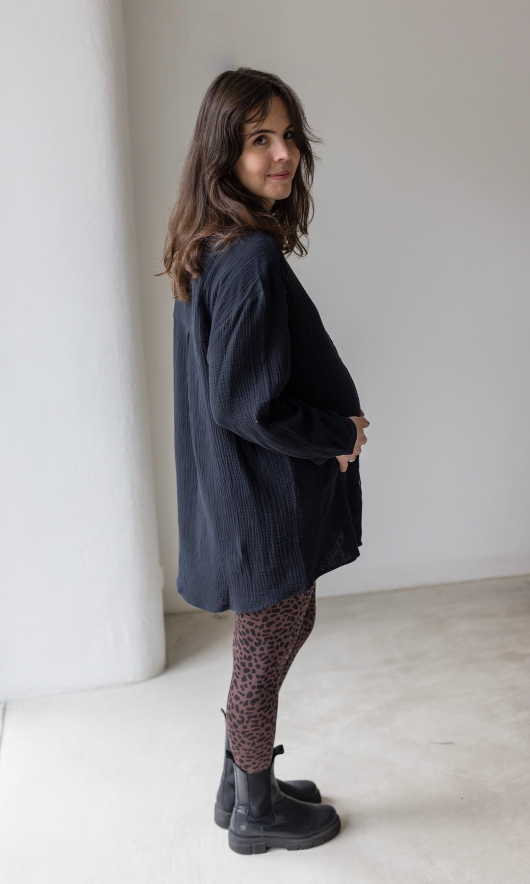 Umstandsleggings braun leo Print mit schwarzem Musselin Hemd schwarzen Stiefeln seitlich vor heller Wand Blick nach vorne