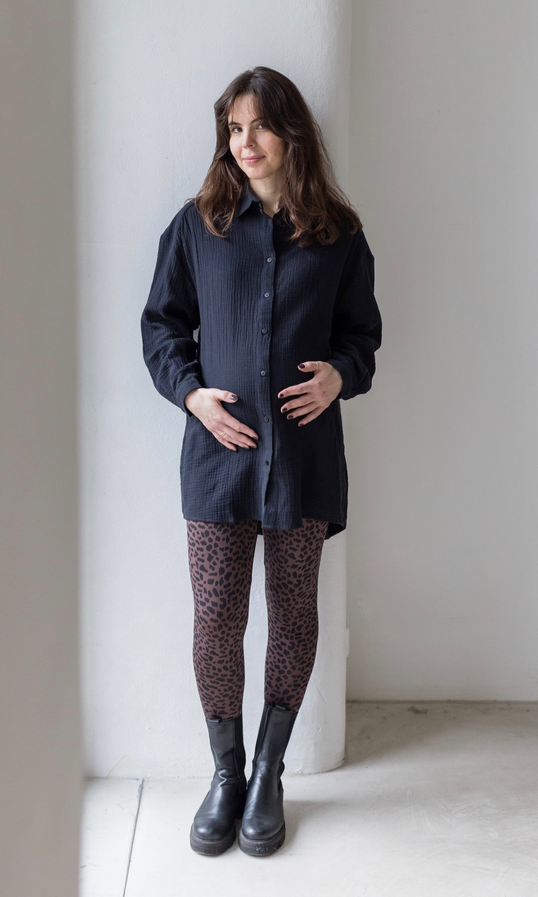 Umstandsleggings braun leo Print mit schwarzem Musselin Hemd schwarzen Stiefeln vor heller Wand Blick nach vorne