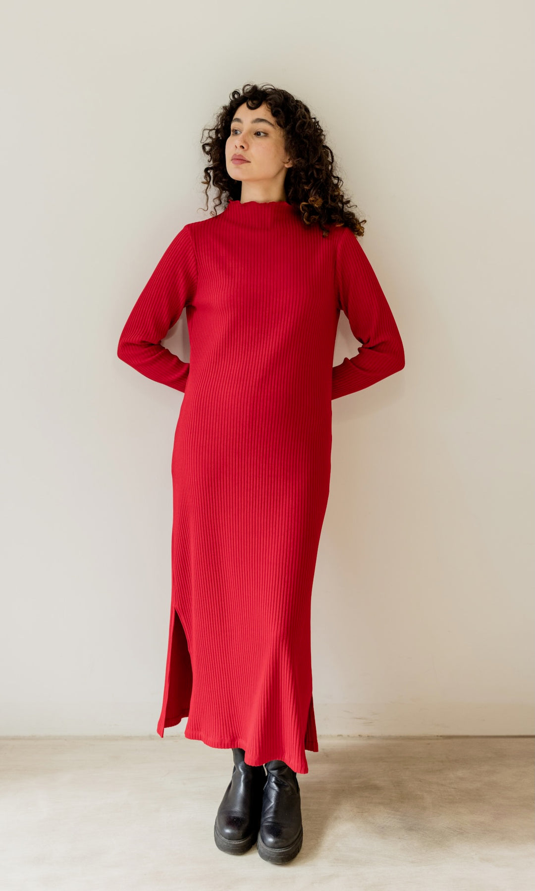 Langes Strickkleid Jil in weinrot mit schwarzen Stiefeln angelehnt an heller Wand Blick seitlich