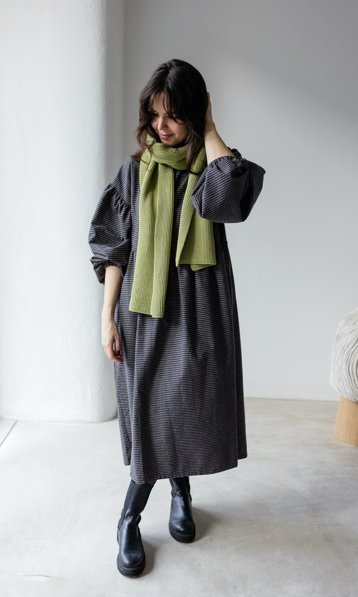 Winter Stillkleid Lovis in karo schwarz mit schwarzen Stiefeln und grünem Schal vor heller Wand Blick nach unten