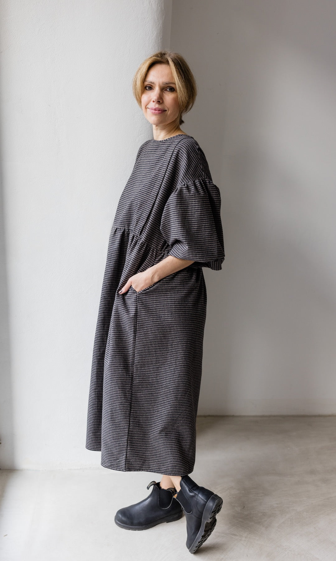 Winter Stillkleid Lovis in karo schwarz mit schwarzen Stiefeln seitlich vor heller Wand Hände in Taschen