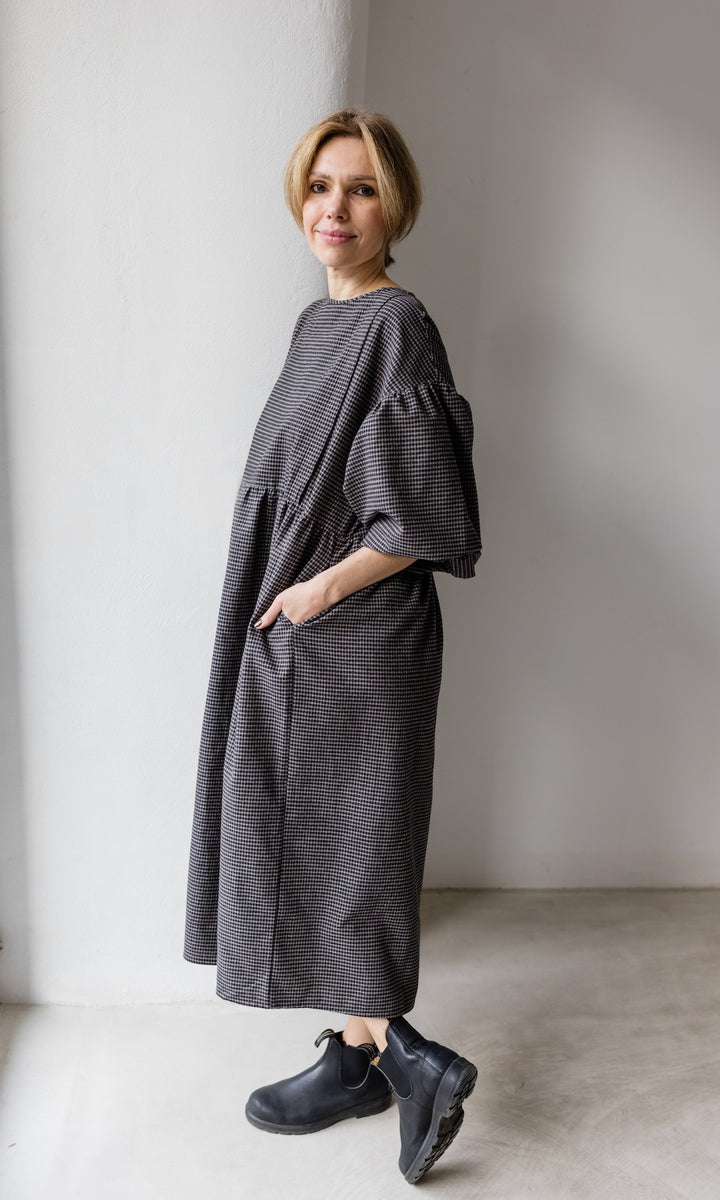 Winter Stillkleid Lovis in karo schwarz mit schwarzen Stiefeln seitlich vor heller Wand Hände in Taschen