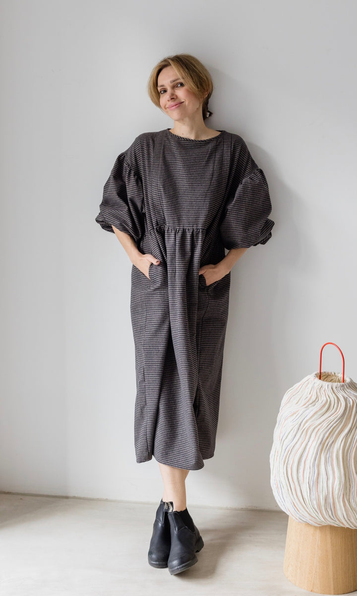 Winter Stillkleid Lovis in karo schwarz mit schwarzen Stiefeln angelehnt an helle Wand Blick nach vorne lächelnd