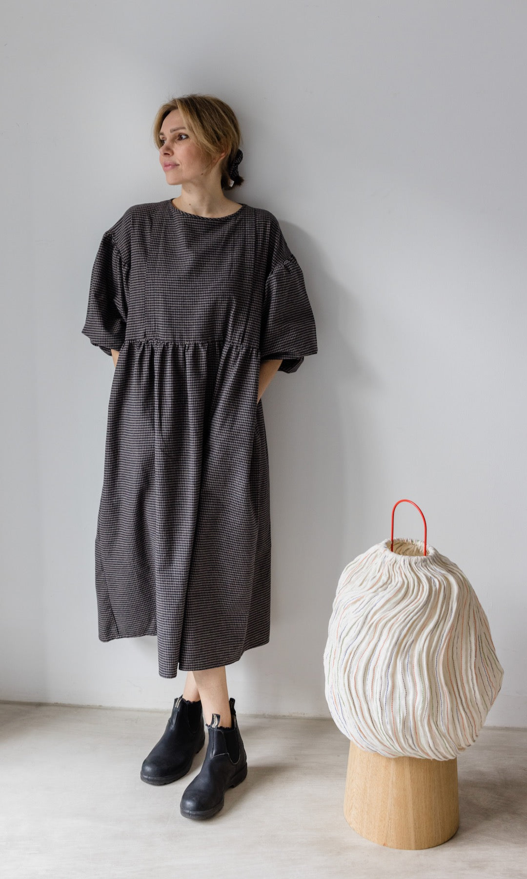 Winter Stillkleid Lovis in karo schwarz mit schwarzen Stiefeln angelehnt an heller Wand Blick seitlich