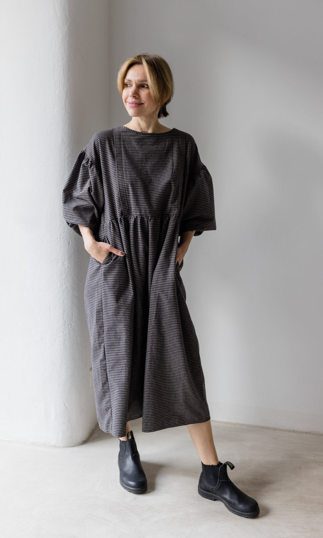 Winter Stillkleid Lovis in karo schwarz mit schwarzen Stiefeln vor heller Wand seitlich Blick seitlich Hände in Taschen