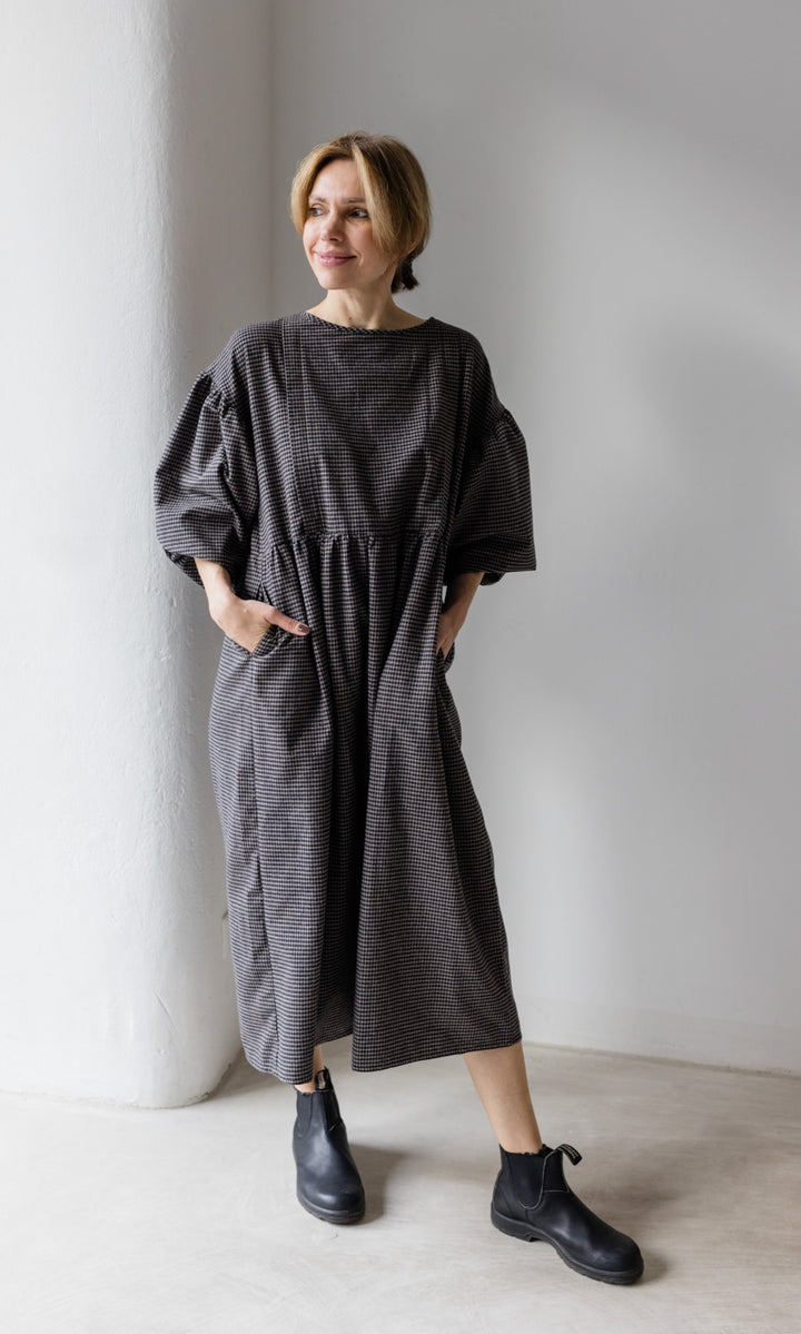 Winter Stillkleid Lovis in karo schwarz mit schwarzen Stiefeln vor heller Wand seitlich Blick seitlich Hände in Taschen