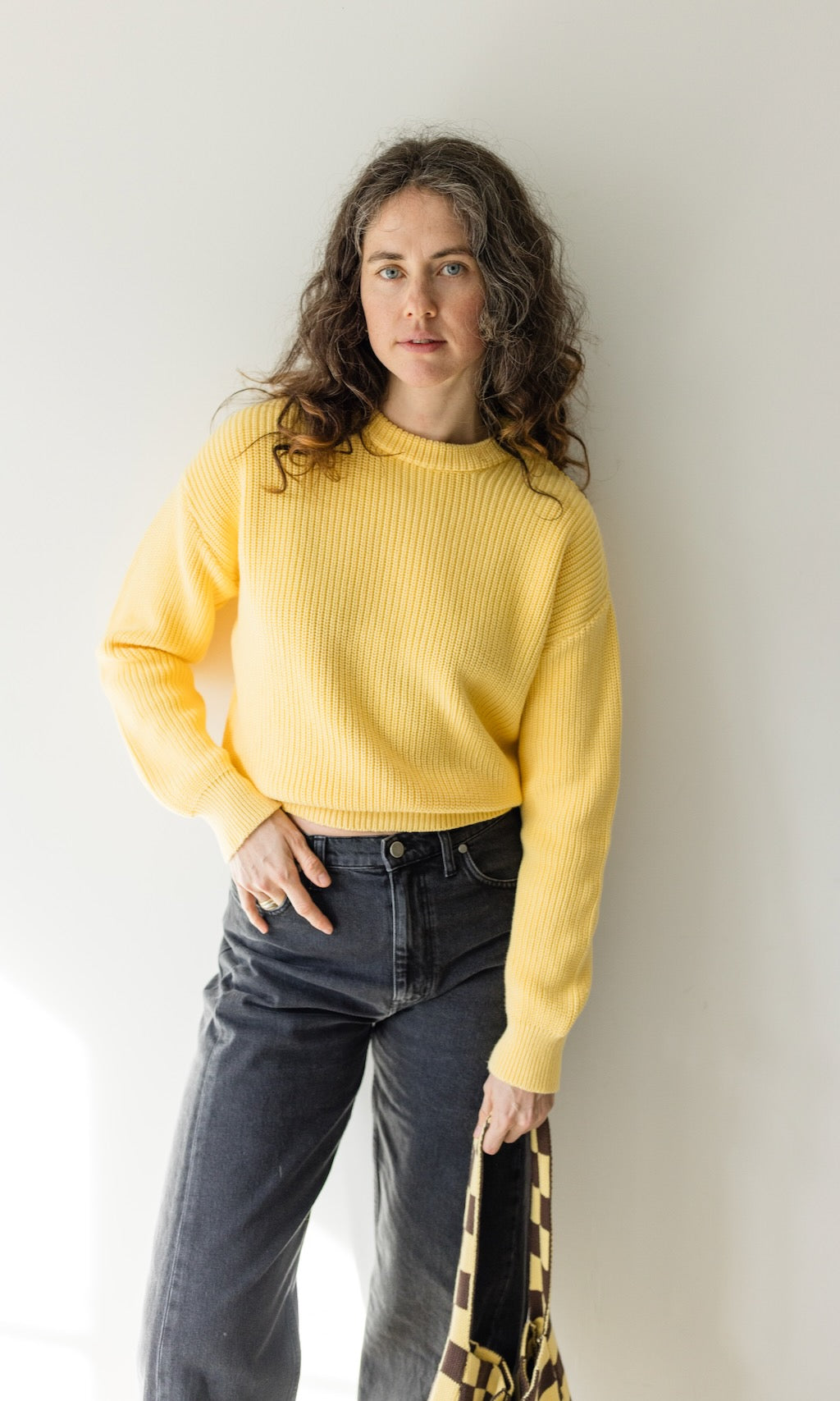 Zitronengelber Strickpullover aus Bio-Baumwolle kombiniert mit schwarzer Barrel-Jeans und gelb/braun-karierter Stricktasche als lässiges Outfit für einen Städtetrip