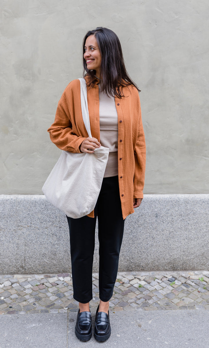 Schwarze Umstandshose im Look mit orangenem Musselin Hemd, hellem Shirt, weißer Mom Bag und schwarzen Schuhen. Ansicht vor heller Hauswand.