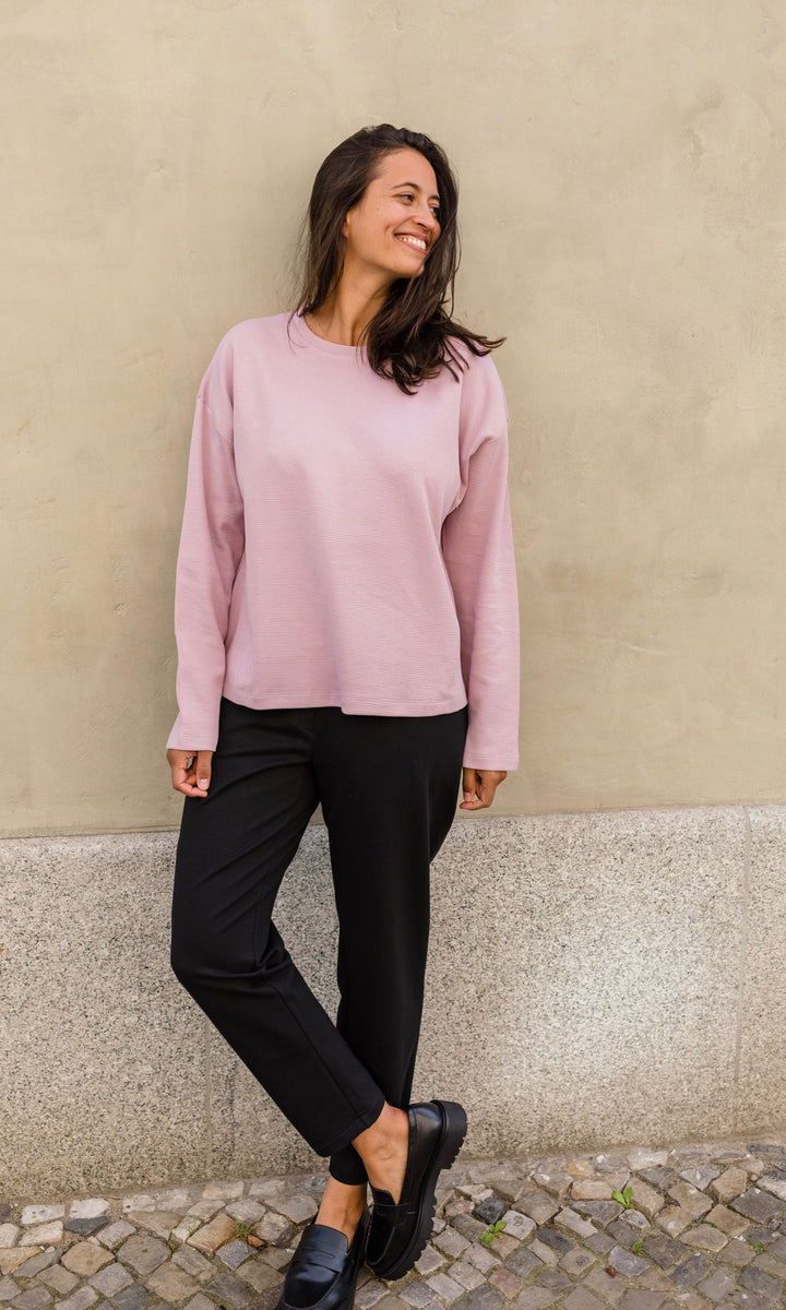 Schwarze Umstandshose im Look mit pinkem Langarmshirt und schwarzen Schuhen. Ansicht vor heller Hauswand.