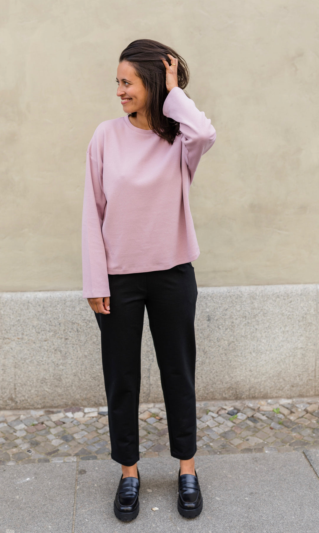 Schwarze Umstandshose im Look mit pinkem Langarmshirt und schwarzen Schuhen. Vorderansicht vor heller Hauswand.