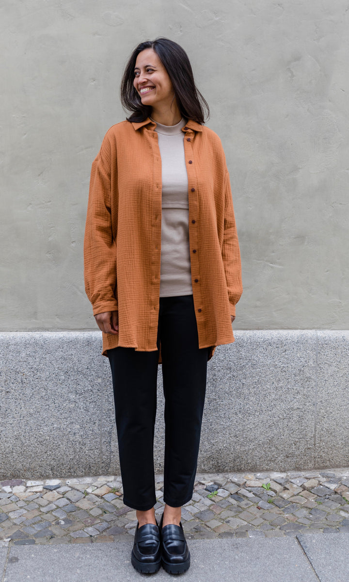 Schwarze Umstandshose im Look mit orangenem Musselin Hemd, hellem Shirt und schwarzen Schuhen. Ansicht vor heller Hauswand.