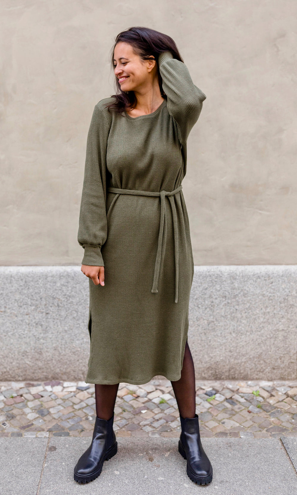 Still Strickkleid Jules in grün olive im festlichen Look mit Strumphose und Stiefeln und Gürtel frontal vor heller Wand Blick seitlich lächelnd