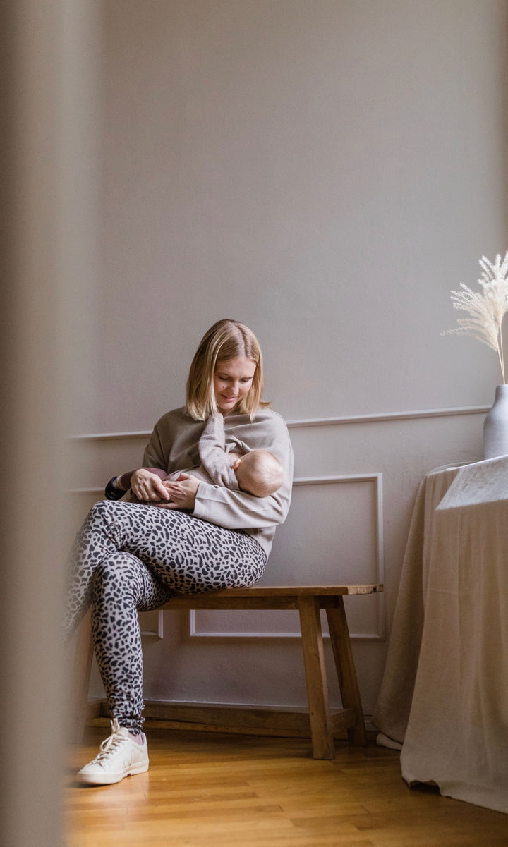 Stillhoodie in sesam weiß sitzend mit Baby im Arm an geöffnetem Reißverschluss mit Leo Leggings Blick auf Kind