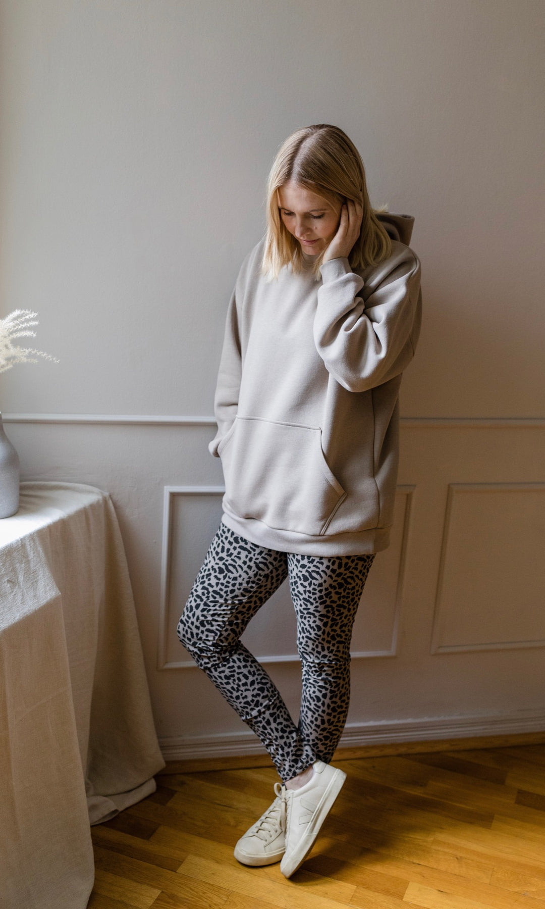 Stillhoodie in sesam weiß stehend mit Leo Leggings neben Tisch Hand in Tasche Blick nach unten