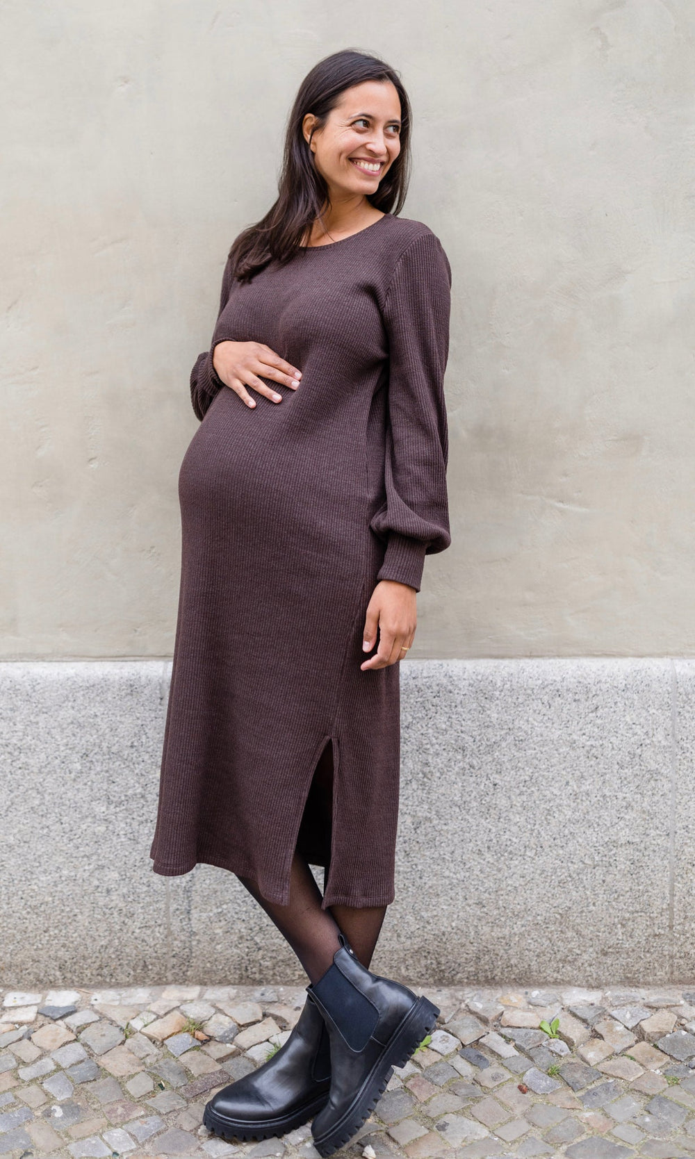 Stillkleid Jules in braunem Strick seitlich im festlichen Look mit Strumpfhose und schwarzen Stiefeln vor Wand Blick seitlich Babybauch mit Hand haltend 