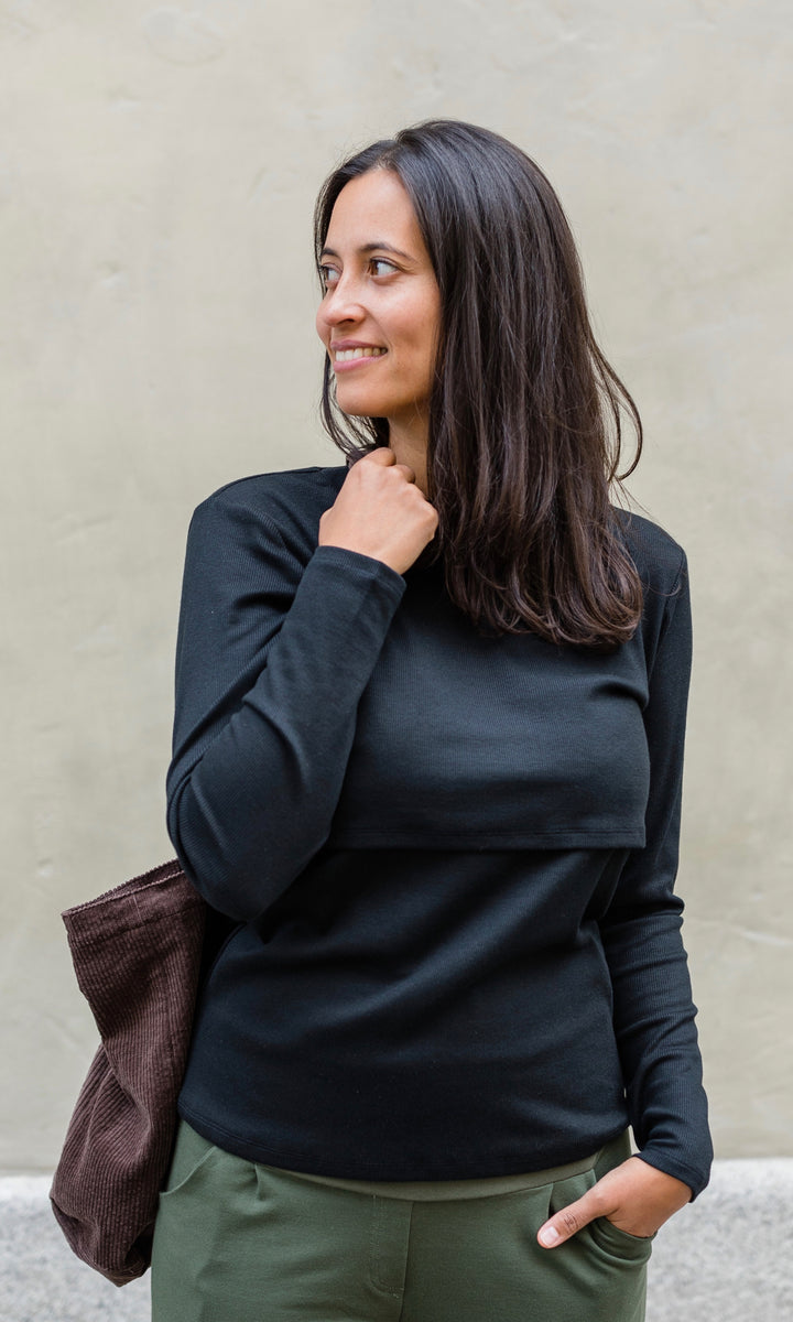 Langarm Stillshirt Cléo mit Rollkragen in schwarz kombiniert mit grüner Hose und brauner Tasche vor grauer Wand im Herbst von vorne Blick seitlich lächelnd