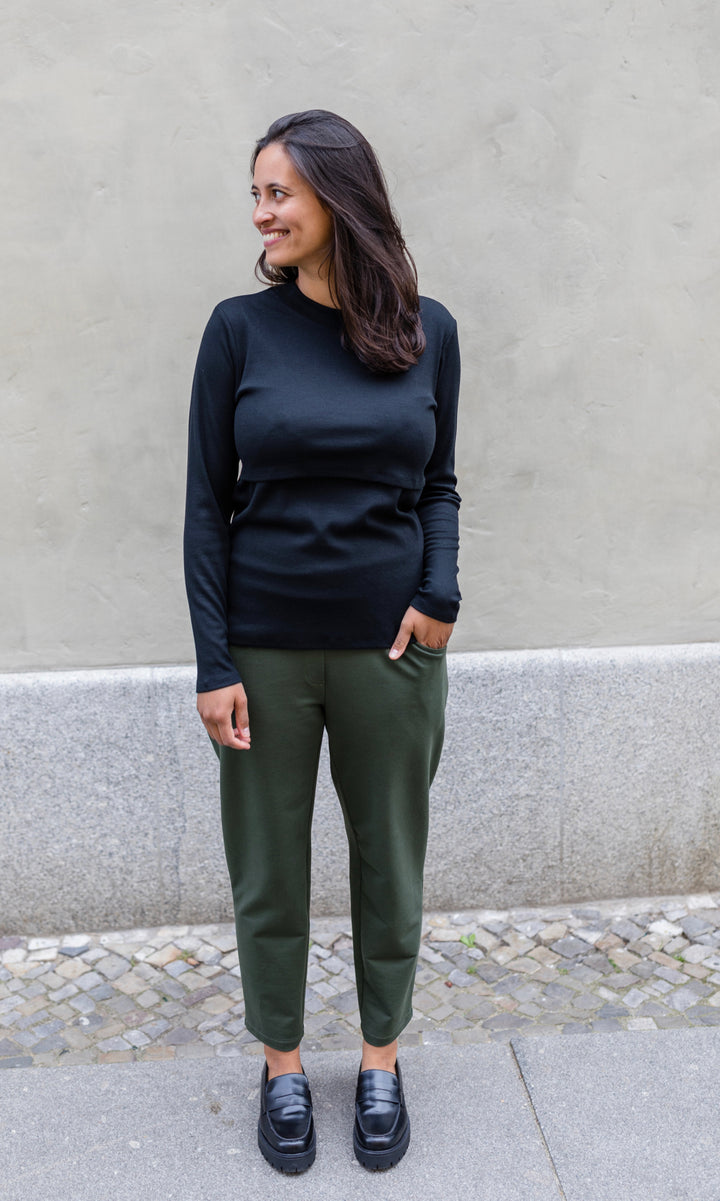 Langarm Stillshirt Cléo mit Rollkragen in schwarz kombiniert mit grüner Hose und schwarzen Schuhen vor grauer Wand im Herbst von vorne Blick seitlich lachend
