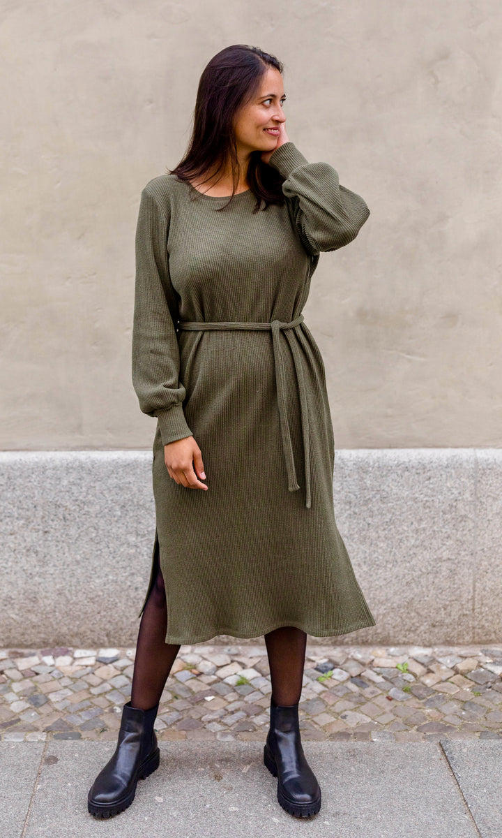 Strickkleid Jules in grün olive mit Stiefeln und Gürtel frontal vor heller Wand Blick seitlich