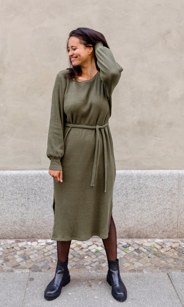 Strickkleid Jules in grün olive mit Gürtel und Stiefeln frontal vor heller Wand Blick seitlich